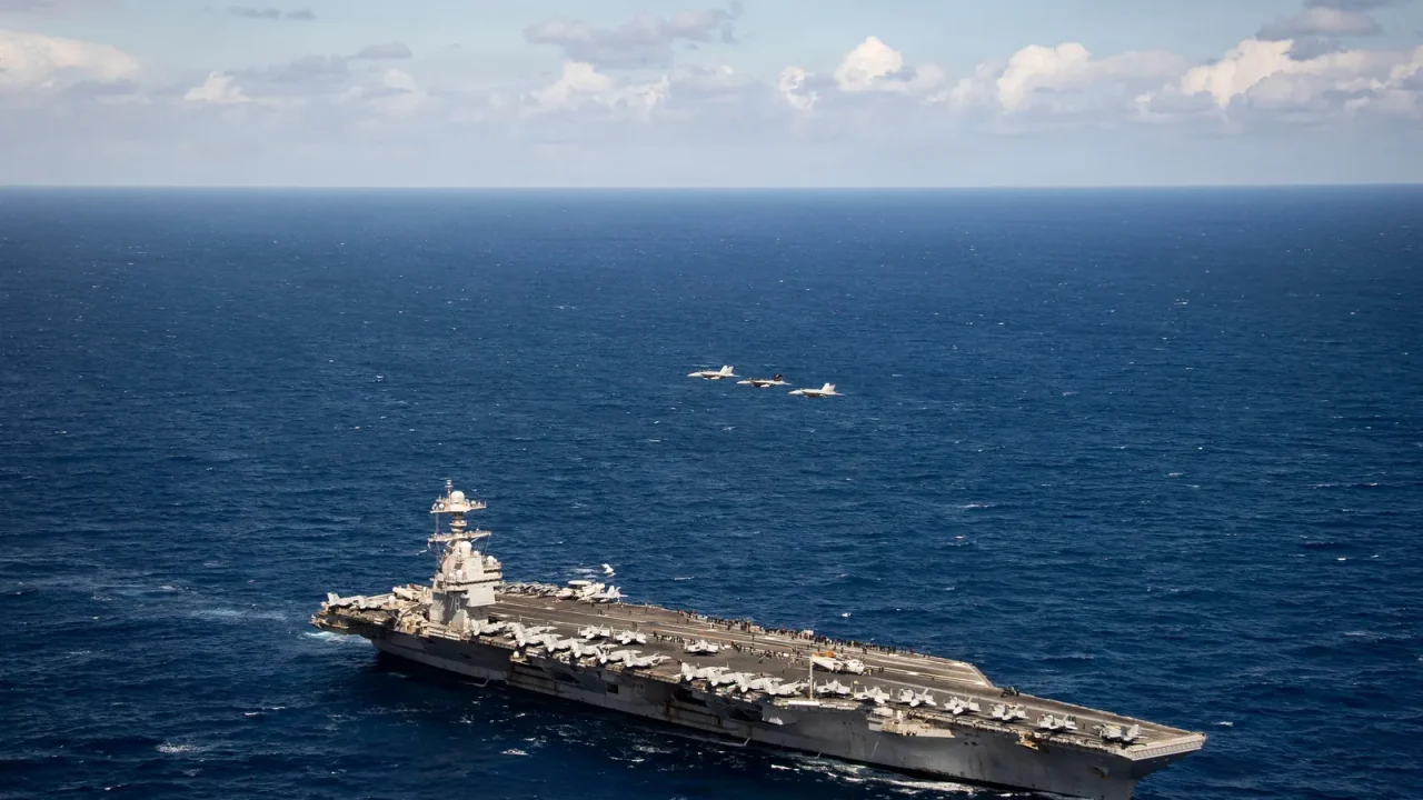 Aircraft carrier USS Gerald R. Ford at sea seen from above, with aircraft parked on the flight deck and jets flying nearby
