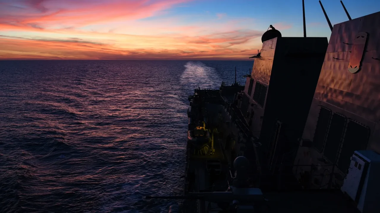 View aft from a Navy destroyer as it leaves a wake across the ocean under a pink and orange sunset
