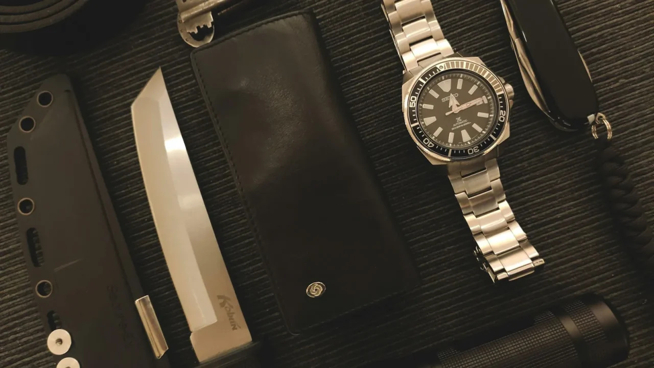 Everyday carry gear laid out on a dark surface, including a dive watch, flashlight, wallet and pocket knife