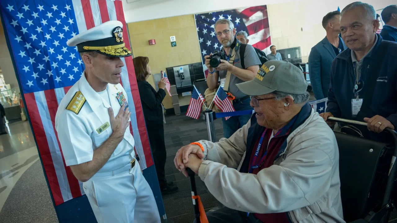 A Navy rear admiral in dress whites greets a Vietnam veteran in a wheelchair at an Honor Flight homecoming, with American flags behind them