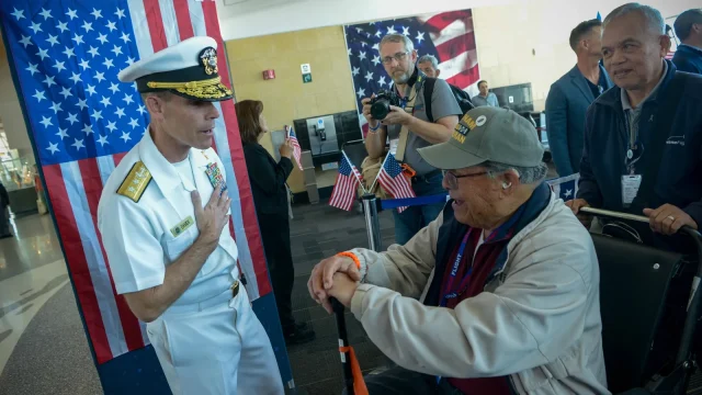 A Navy rear admiral in dress whites greets a Vietnam veteran in a wheelchair at an Honor Flight homecoming, with American flags behind them