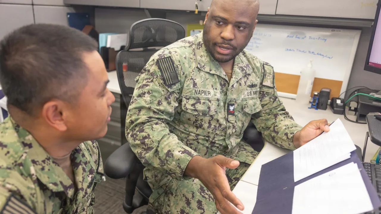 A Navy yeoman at a desk goes through paperwork with another sailor at an administrative office