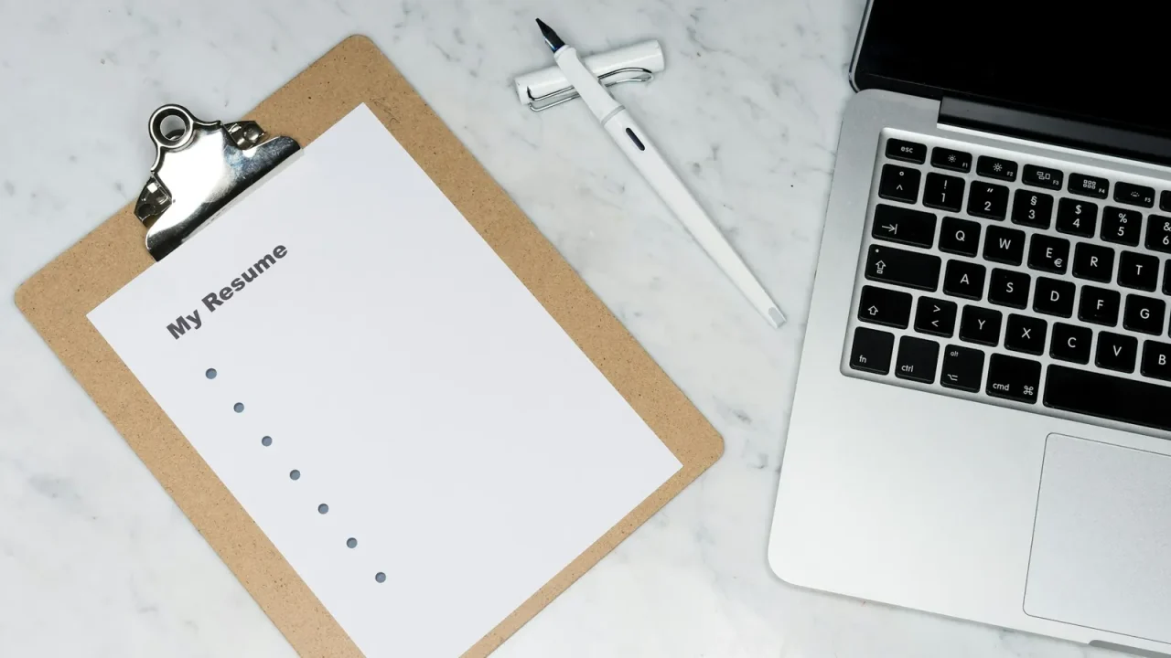 Clipboard holding a printed resume beside a laptop on a marble desk