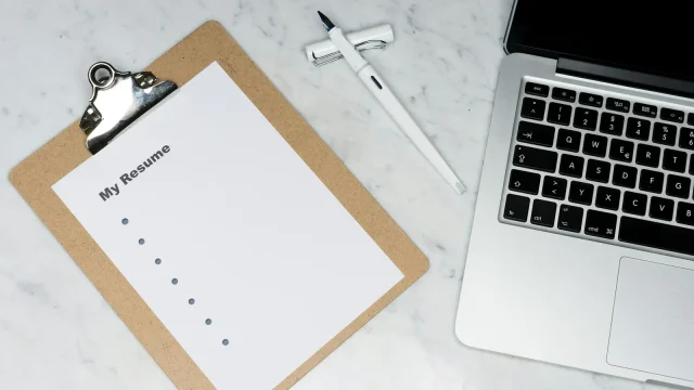 Clipboard holding a printed resume beside a laptop on a marble desk
