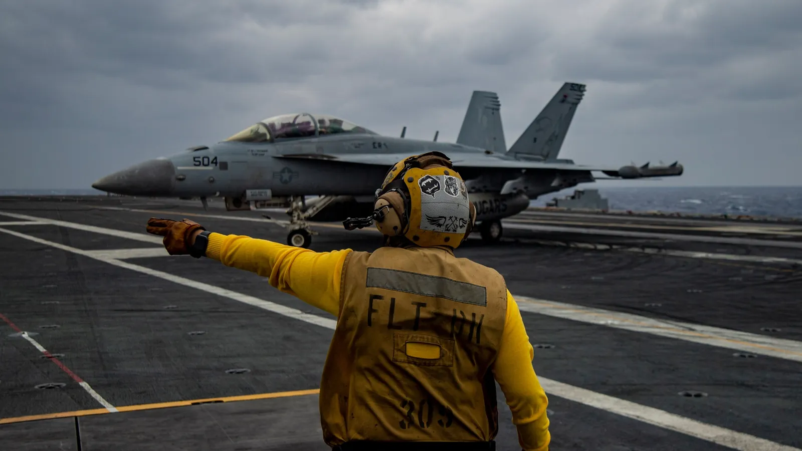 Sailor in a yellow jersey directs a jet on the flight deck of an aircraft carrier