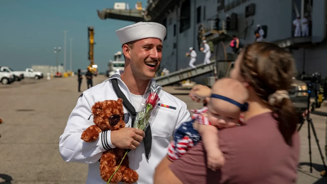 A Navy sailor in dress whites holding a teddy bear and a rose smiles at his wife and baby daughter on the pier at a ship's homecoming