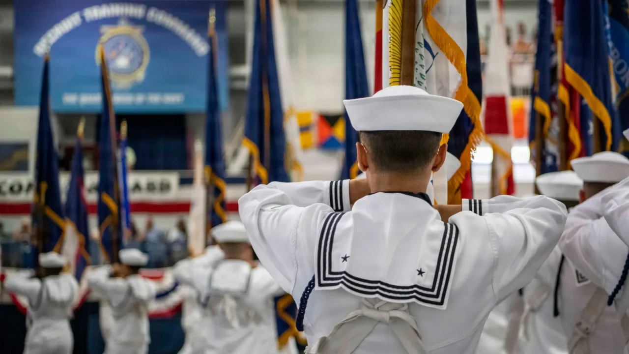 New sailor in dress whites salutes toward a row of flags during a boot camp graduation ceremony