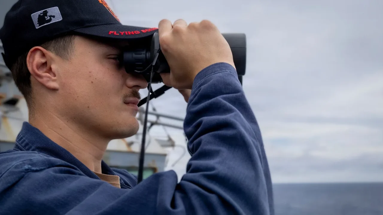 Sailor looks through binoculars while standing watch on the bridge wing of a destroyer