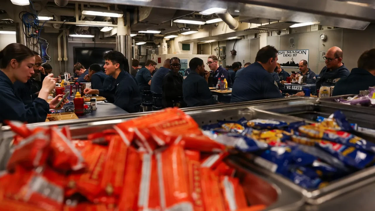 Sailors eat a meal at crowded tables on the mess deck of a Navy destroyer