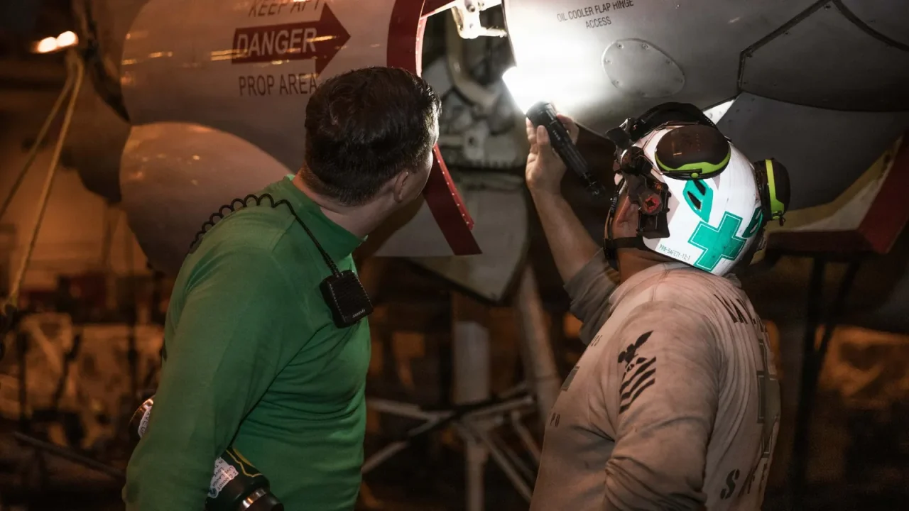 Two Navy sailors use a torch to inspect the landing gear of an aircraft in the hangar bay of an aircraft carrier