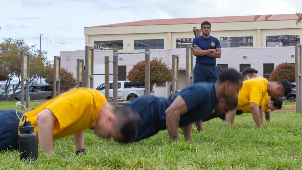 Sailors perform push-ups on a grass field while a fitness leader observes during a physical readiness test