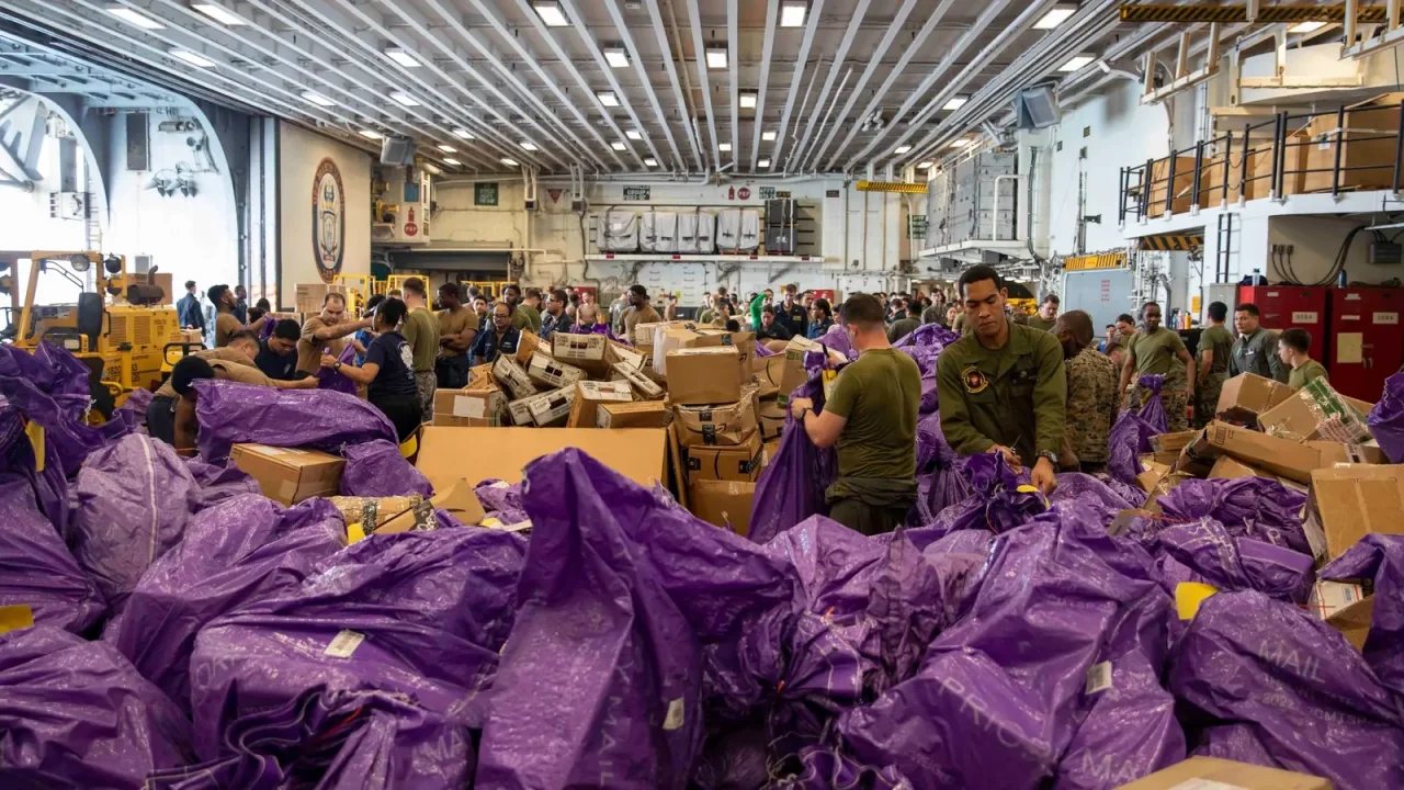 Sailors and Marines sort stacks of boxes and mail bags in a ship's hangar bay during mail call