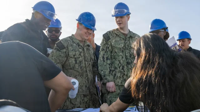 Navy sailors in hard hats talk to community college representatives at an education fair on the pier
