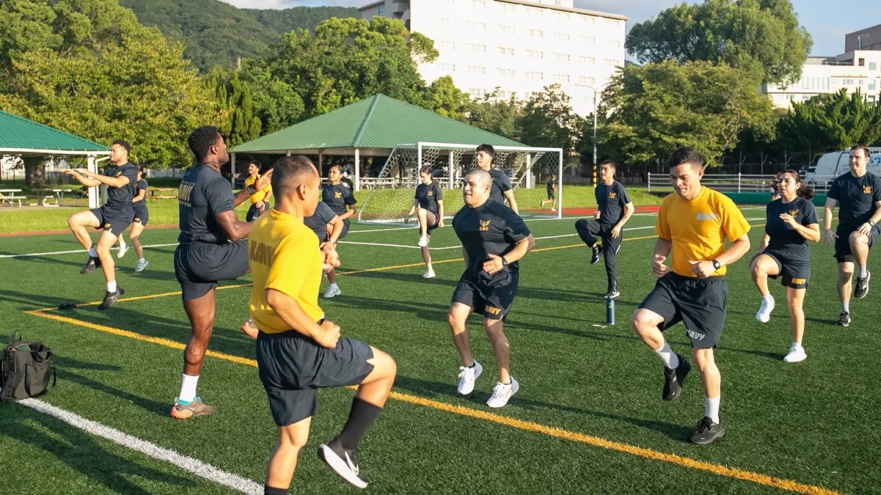 Sailors in Navy physical training uniforms warm up together on an athletic field before a fitness test