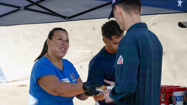 A USO volunteer in a blue shirt hands breakfast to Navy sailors under a canopy on the pier