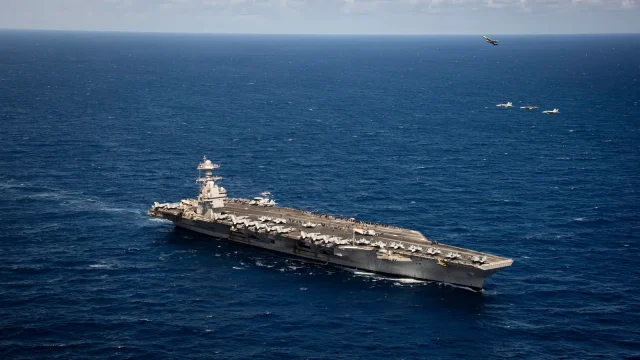 Aerial view of the aircraft carrier USS Gerald R. Ford underway as four fighter jets fly over it in formation